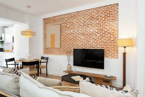 A modern living room with a brick wall, a flat-screen TV, a sofa, a coffee table, and a dining area in the background. - Beautiful one-bedroom apartment on Calle Castillo, Madrid, perfect for extended stays. Mediterranean style blends seamlessly with rustic touches of exposed brick and natural wood, creating a warm, bright, and inviting atmosphere. (MADRID)