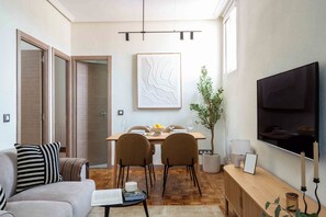 A modern living room with a sofa, a dining table set for two, a television mounted on the wall, and a large window providing natural light. - Private and quiet on Cuesta de Santo Domingo, this three bedroom apartment sits in one of the best locations in Madrid, with the Royal Palace and Teatro Real just steps away and Puerta del Sol and Plaza Mayor an easy walk beyond. (MADRID)