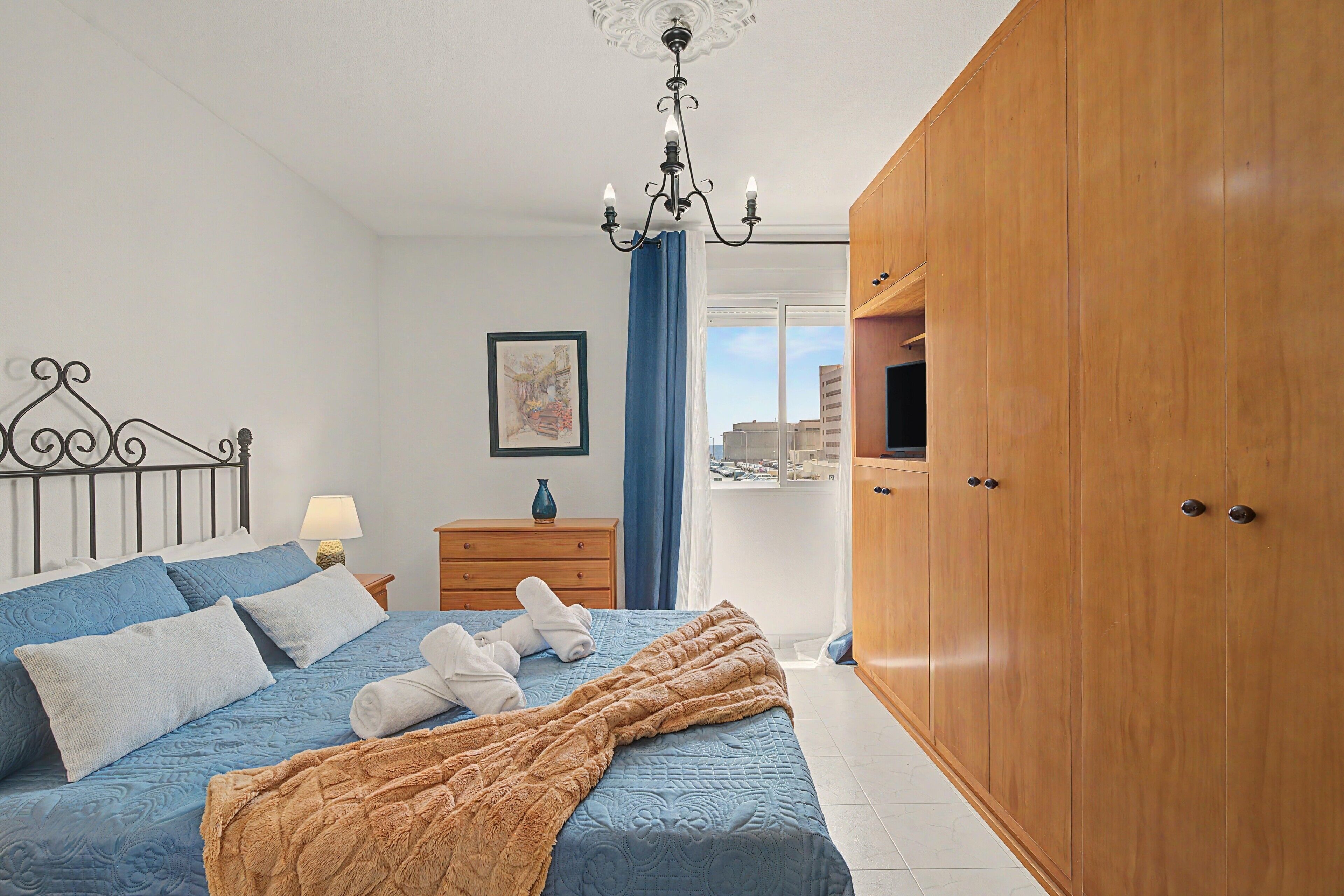 A bedroom with a bed, a wooden wardrobe, a nightstand, a lamp, a framed picture, and a view through a window.