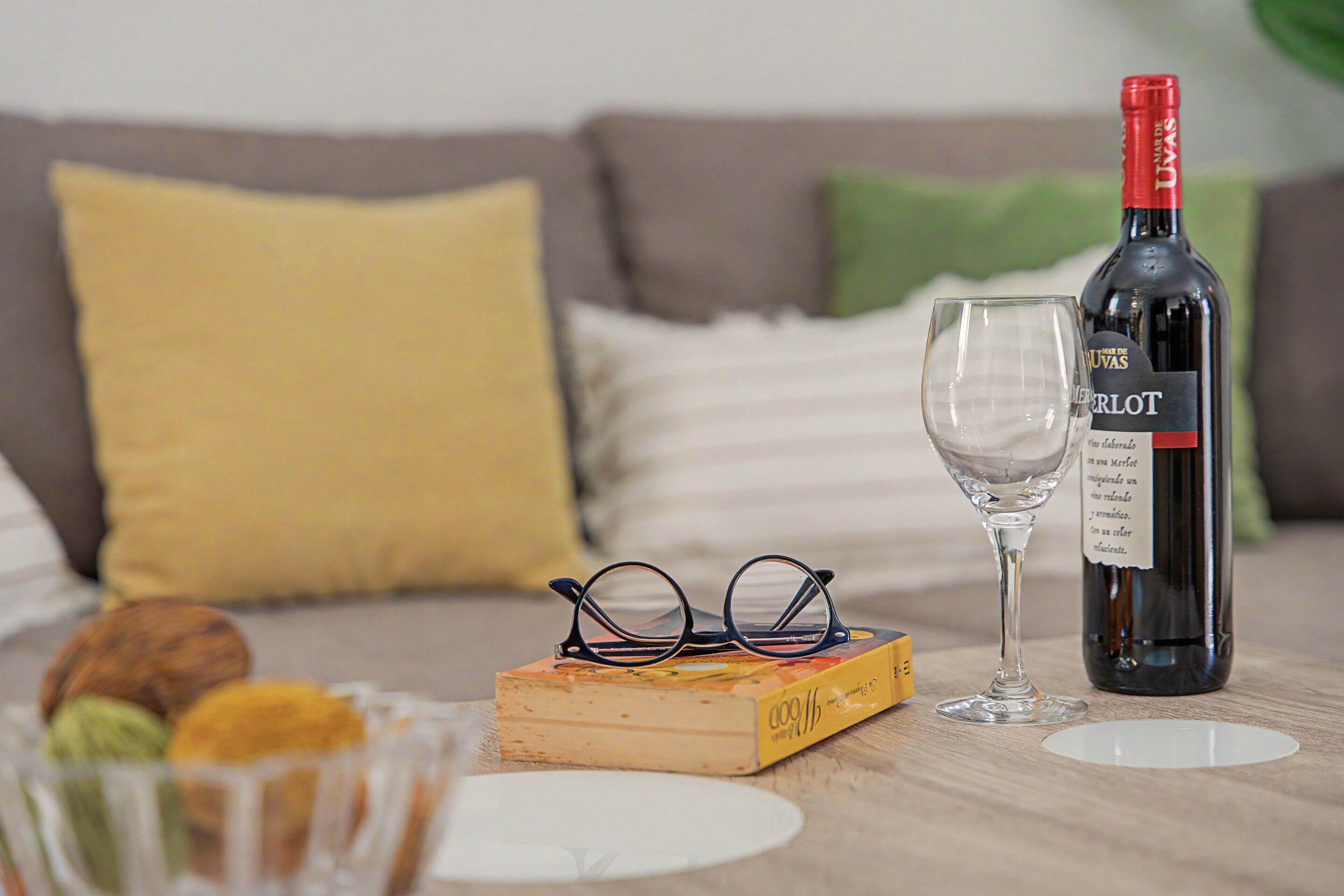 Een fles Merlot wijn, een wijnglas, een paar brillen en een boek op een houten tafel.