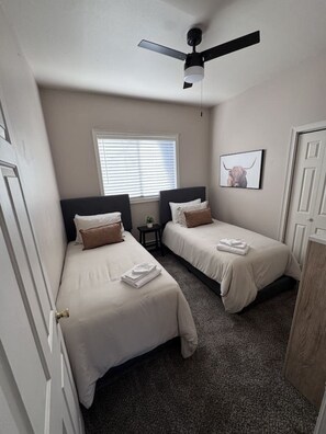 A hotel room with two beds, a ceiling fan, a window with blinds, and a framed picture on the wall.