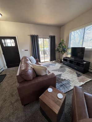 A living room with a brown leather sofa, a wooden coffee table, a television, and a large window with blinds.