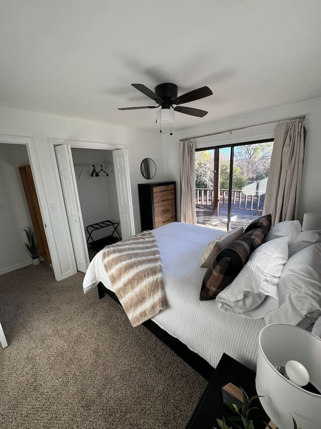 Um quarto com cama, ventilador de teto, janela com cortinas e vista para o exterior.