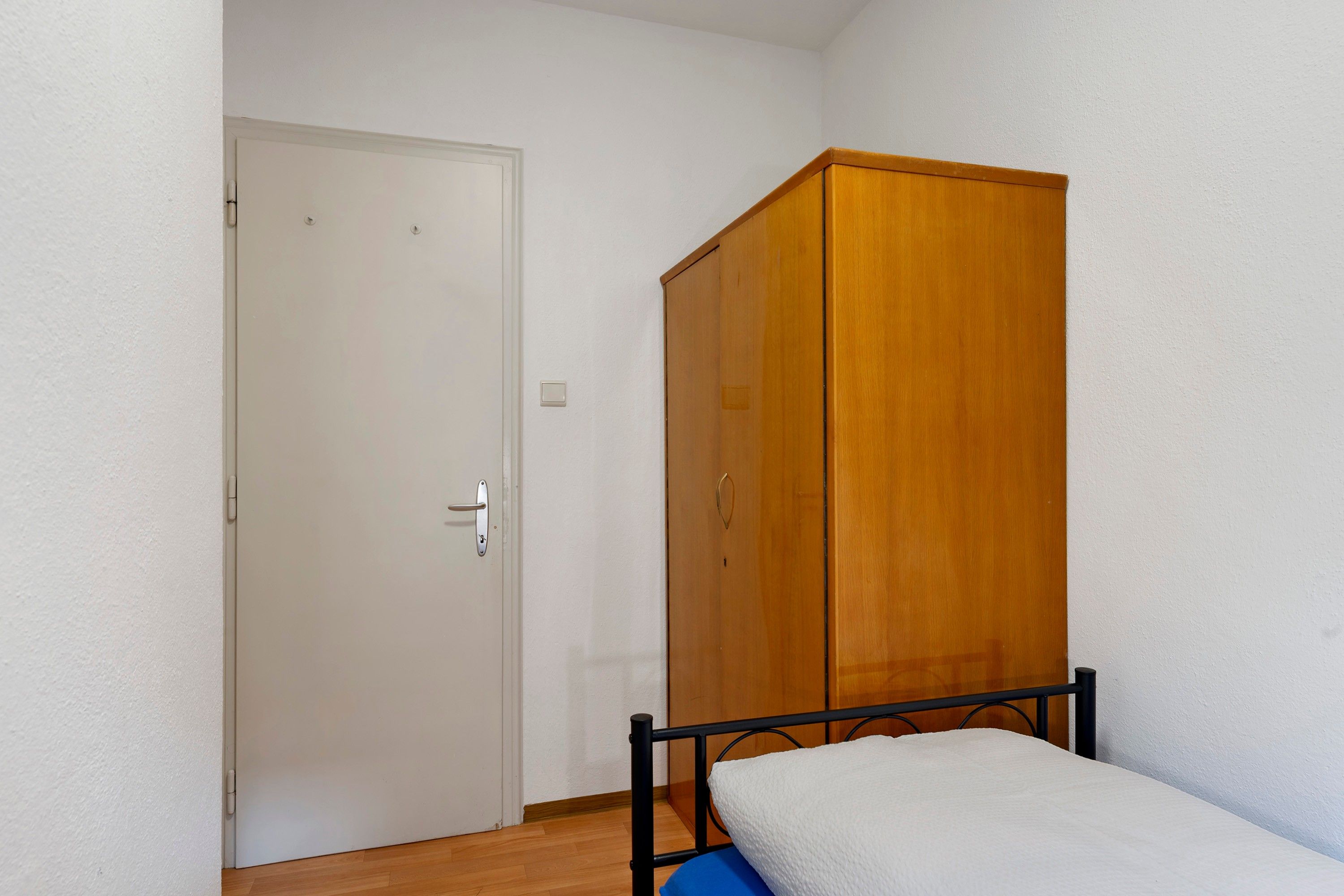 Quarto pequeno com guarda-roupa de madeira, porta branca e estrutura de cama de metal.