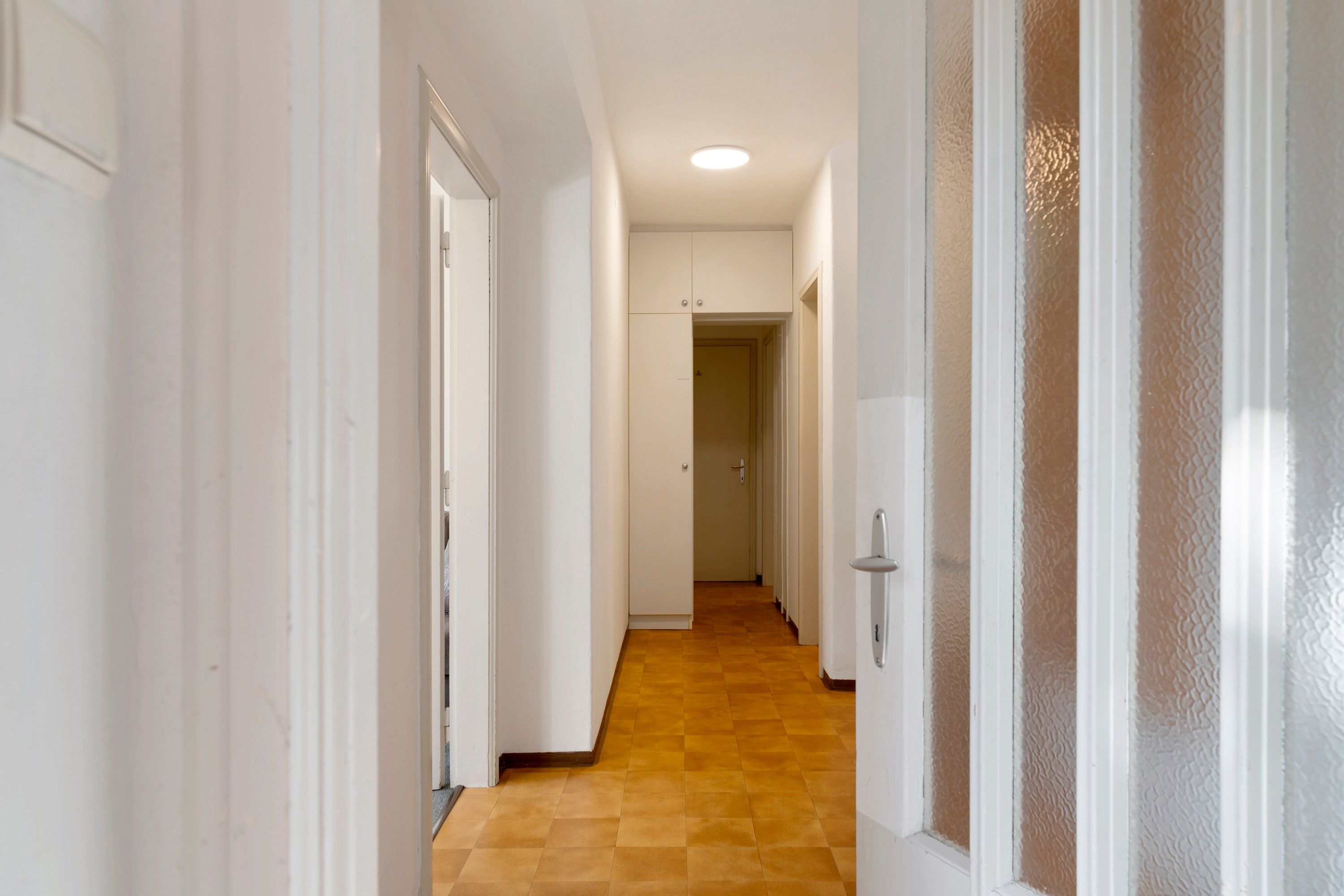 Un couloir aux murs blancs, doté d’un plancher en bois et d’une porte en verre dépoli.
