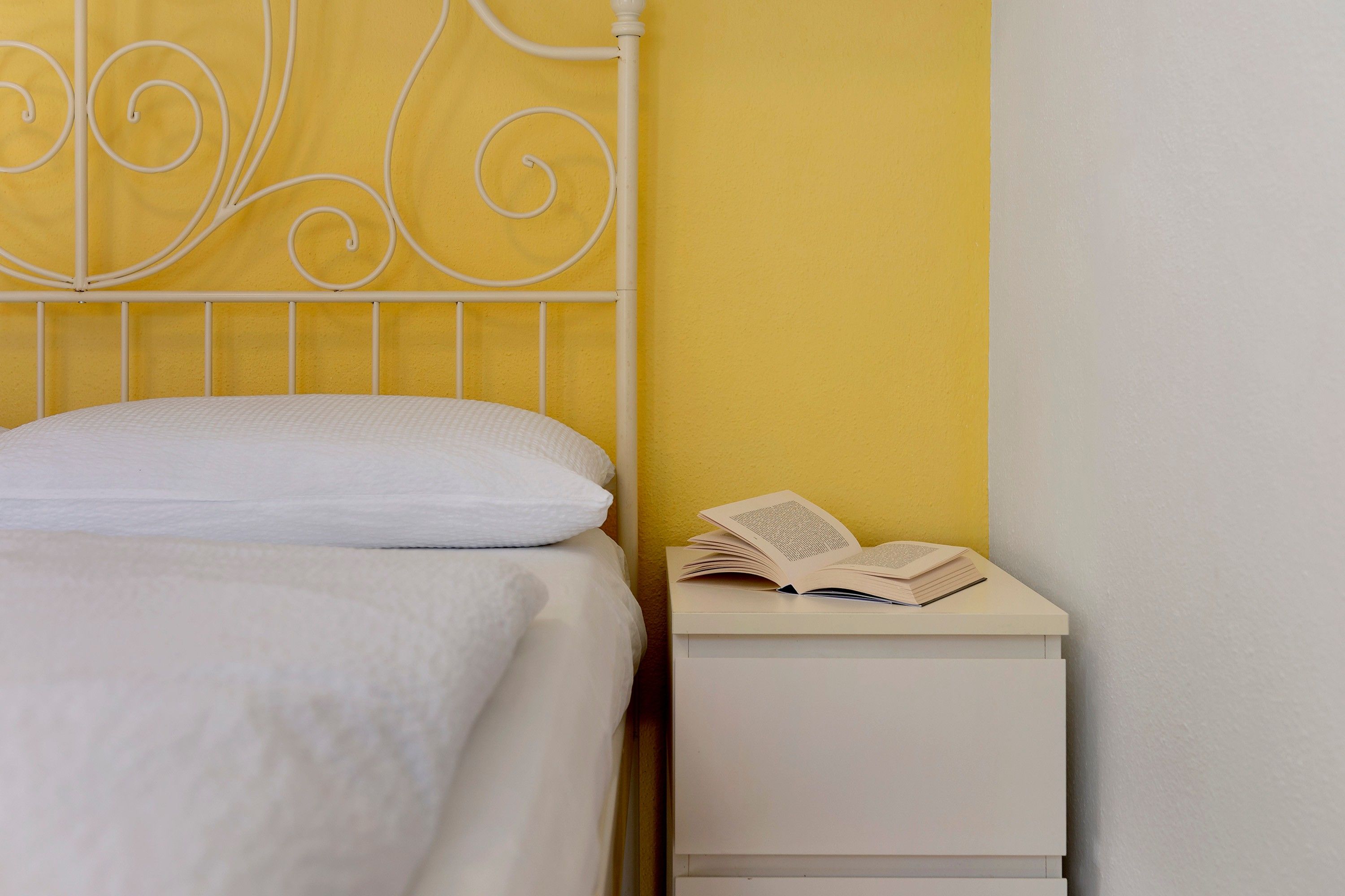 Un lit avec une literie blanche et un oreiller blanc, une table de chevet blanche avec un livre et un stylo, et un mur jaune.