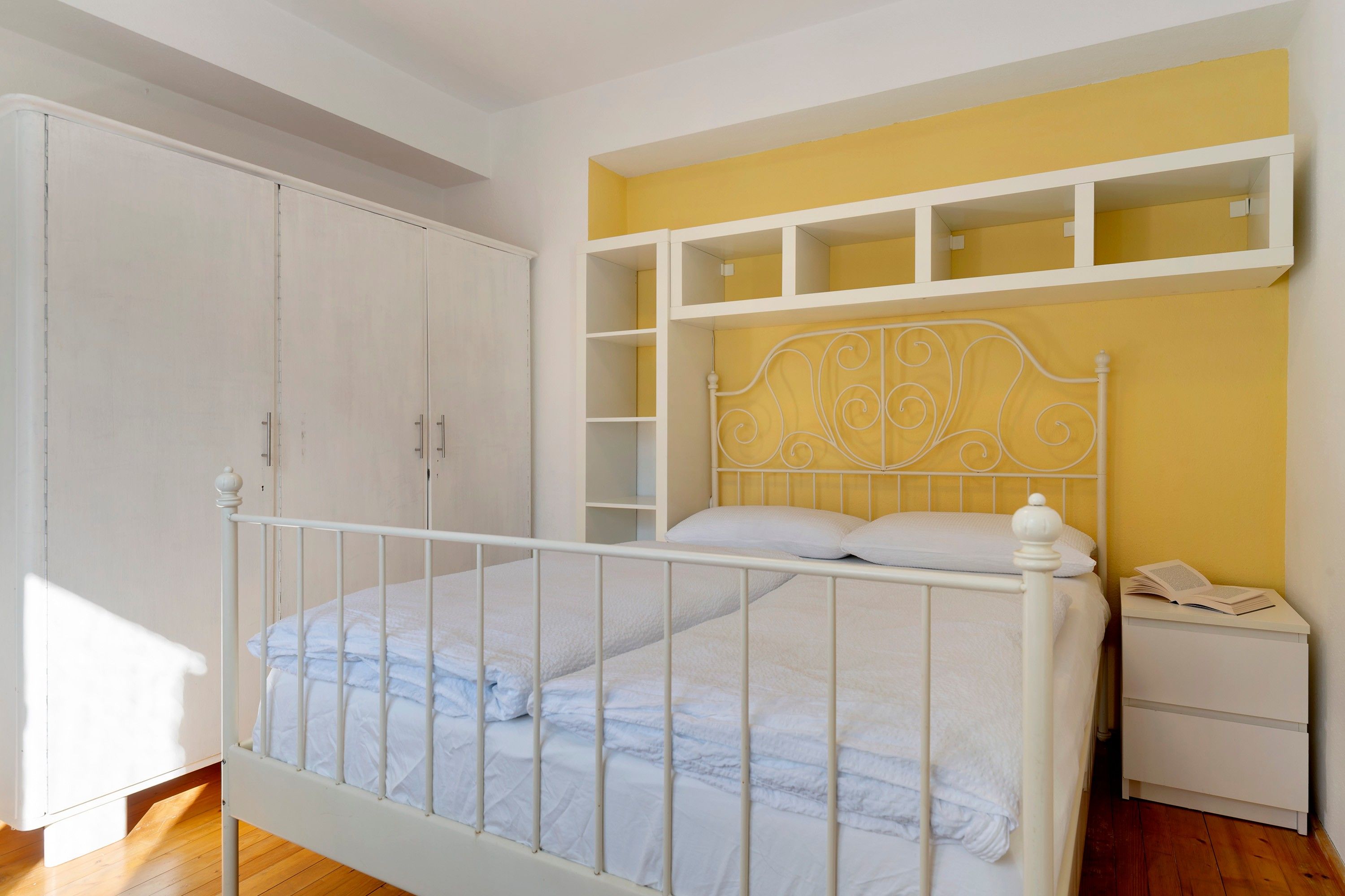 Un dormitorio con estructura de cama metálica blanca, mesita de noche blanca y armario.