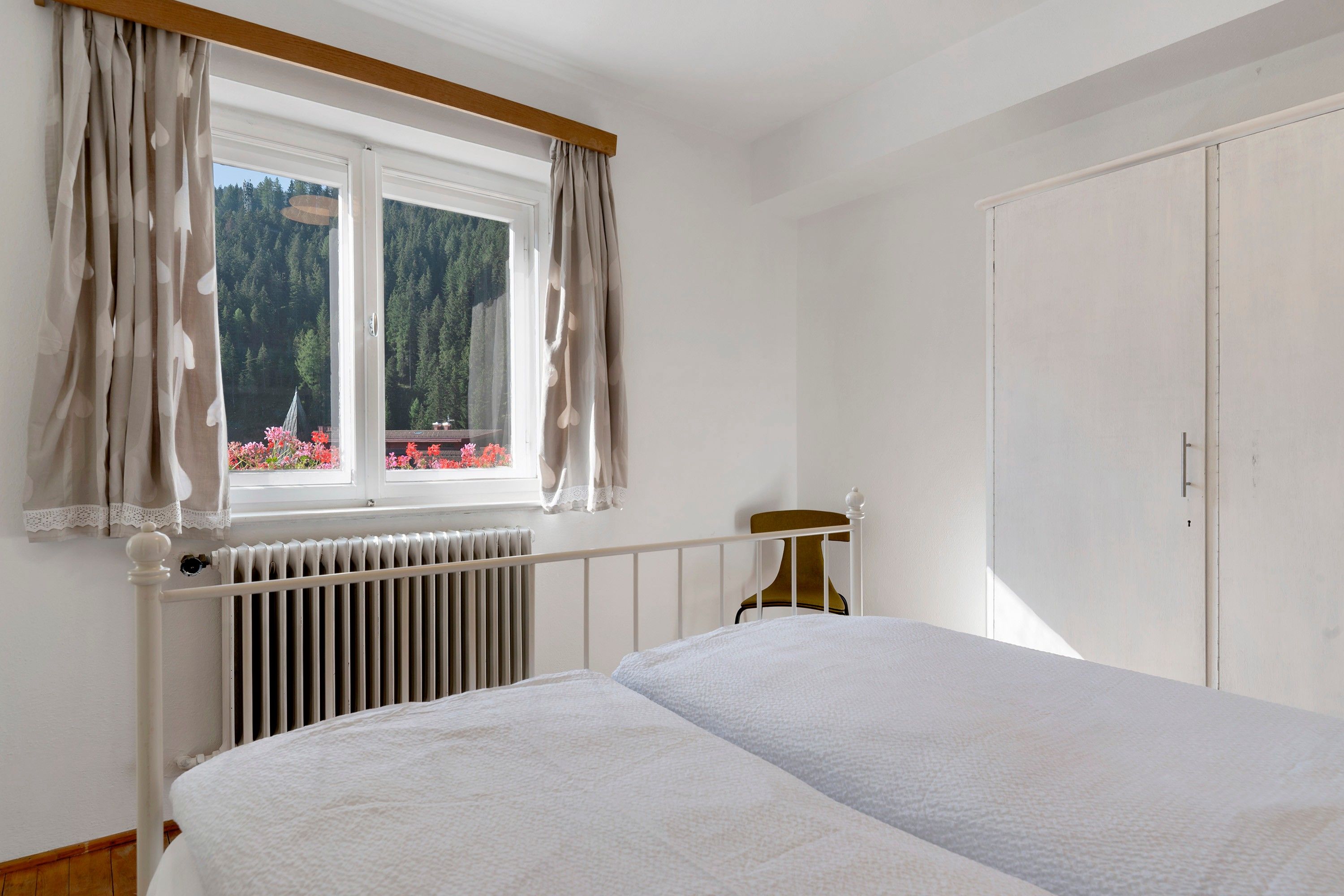 A bedroom with a bed, a radiator, a window with curtains, and a view of a forest.