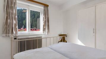 A bedroom with a bed, a radiator, a window with curtains, and a view of a forest.