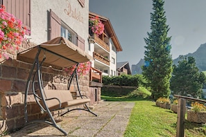 Un bâtiment doté d’une façade en pierre, d’un balcon en bois orné de jardinières et offrant une vue dégagée sur une montagne en arrière-plan.