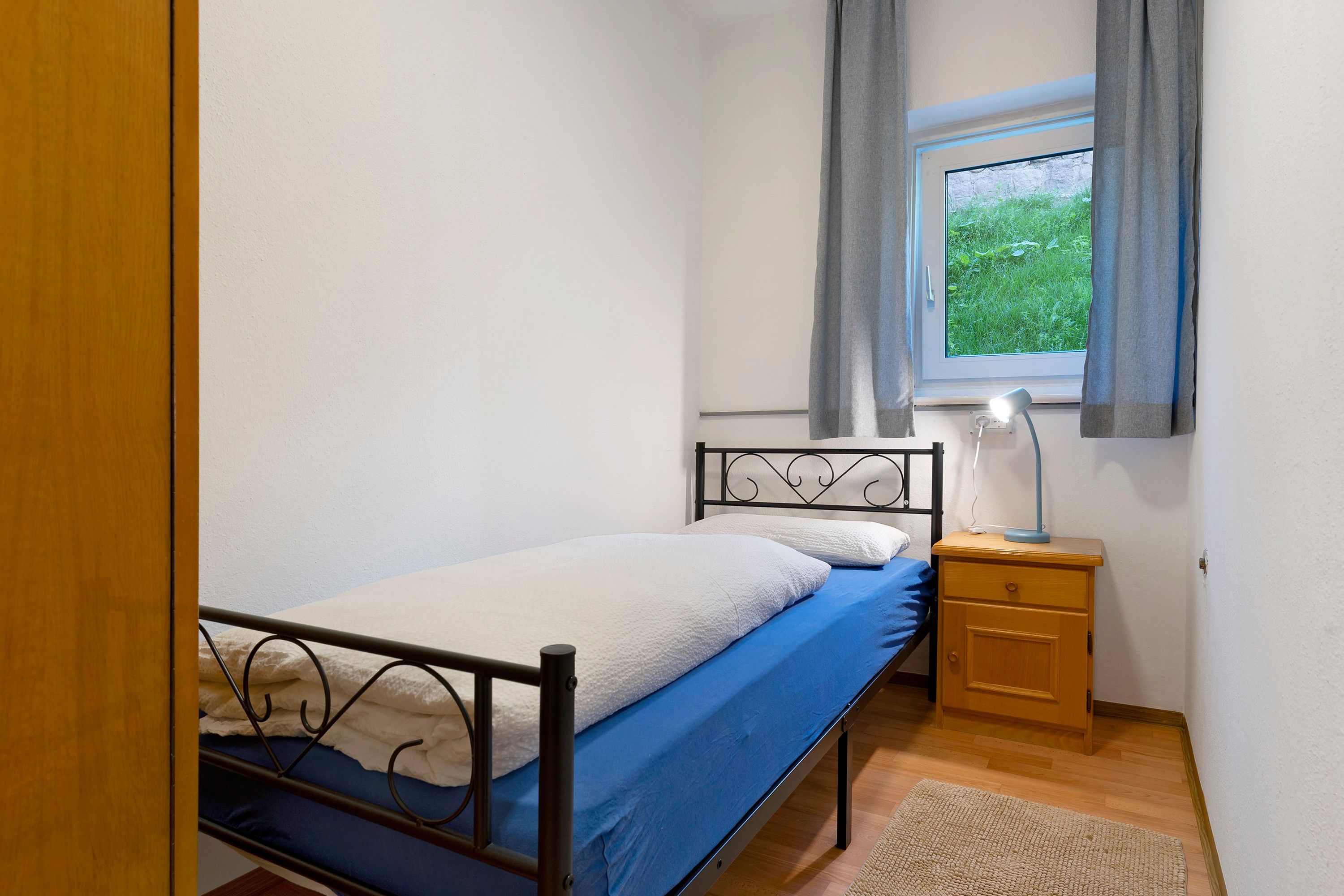 A small, single-bed room with a wooden nightstand, a bedside lamp, and a window with curtains.