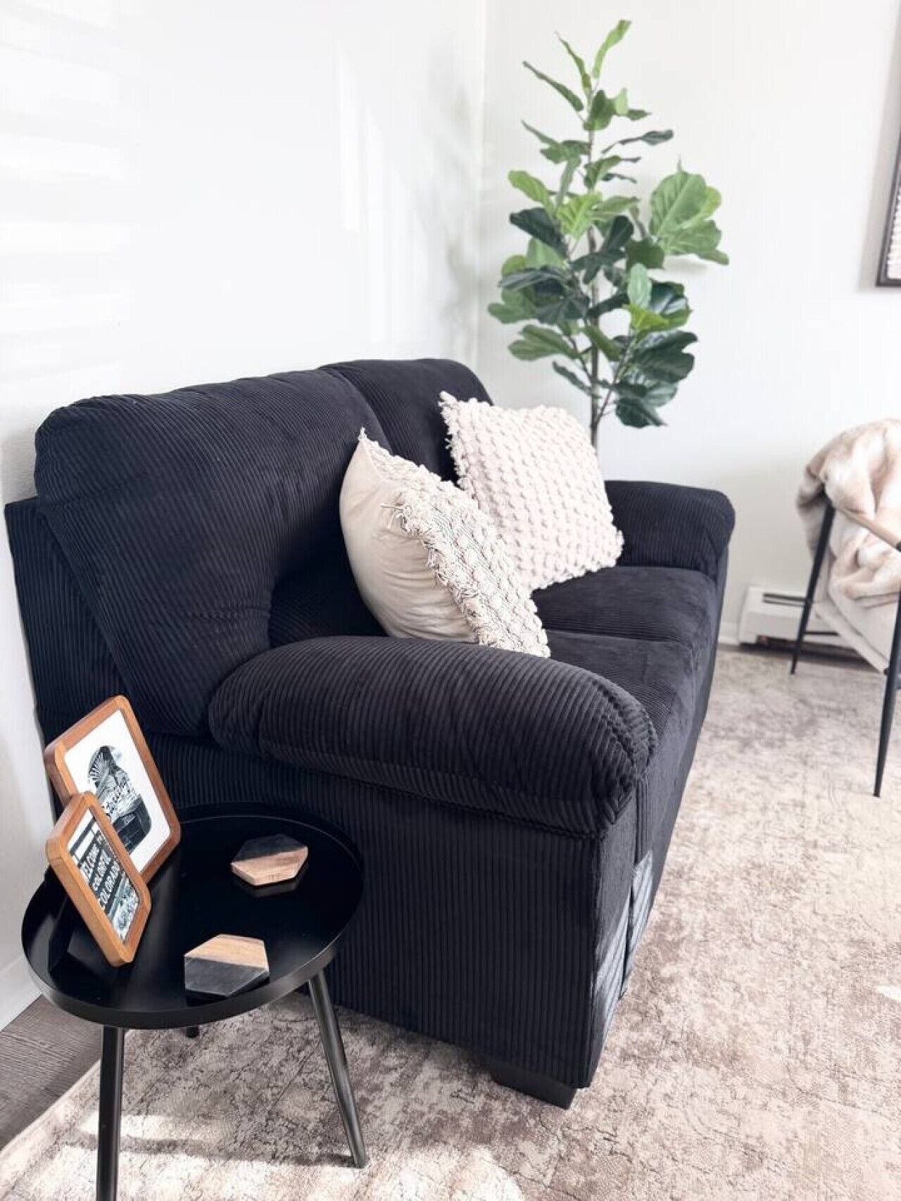 Um sofá preto com uma almofada texturizada, uma pequena mesa lateral preta com um quadro emoldurado e um bloco de madeira, e uma grande planta verde ao fundo.