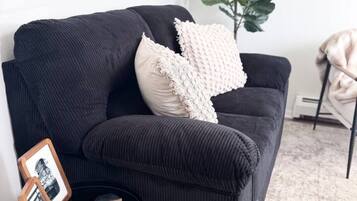 Um sofá preto com uma almofada texturizada, uma pequena mesa lateral preta com um quadro emoldurado e um bloco de madeira, e uma grande planta verde ao fundo.