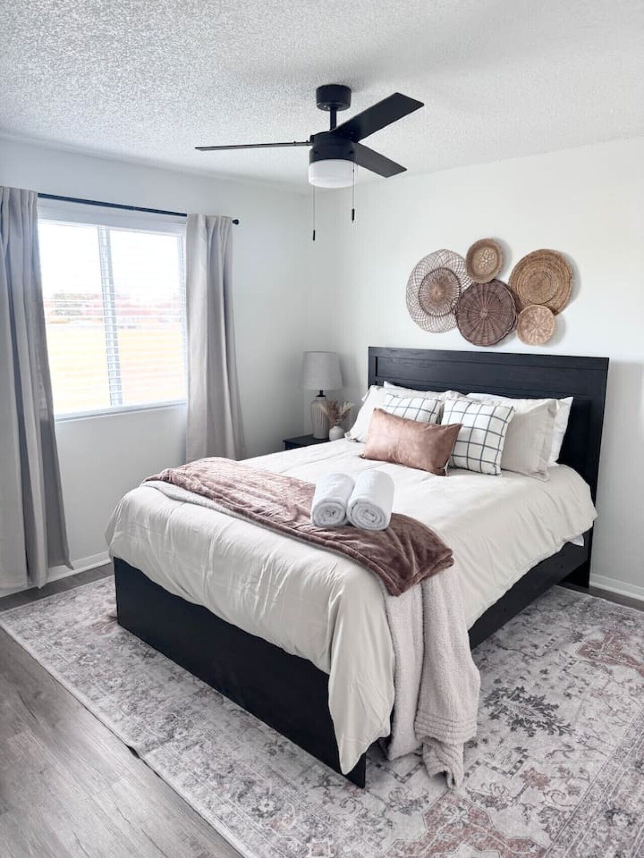 Un dormitorio con cama, ventilador de techo, ventana con cortinas, mesita de noche con lámpara y decoración mural.