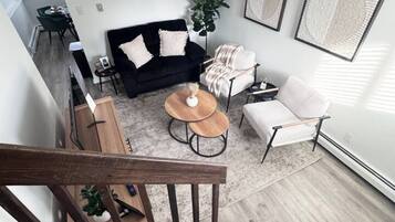 Uma sala de estar moderna com um sofá preto, uma poltrona branca, uma mesa de centro e uma escadaria que leva a outro andar.