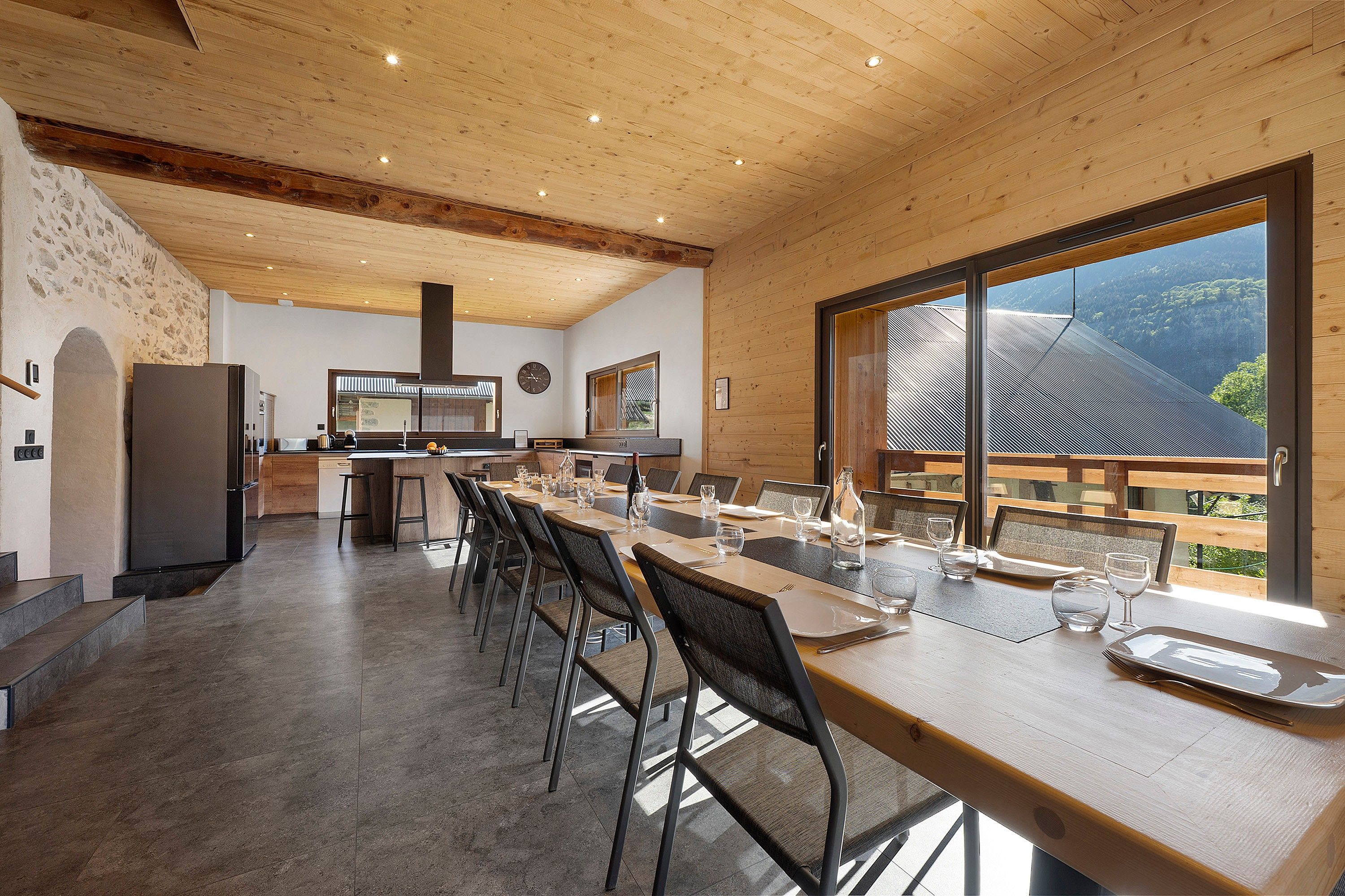 A modern kitchen with a large island, wooden ceiling, and a view of a forest through a large window.