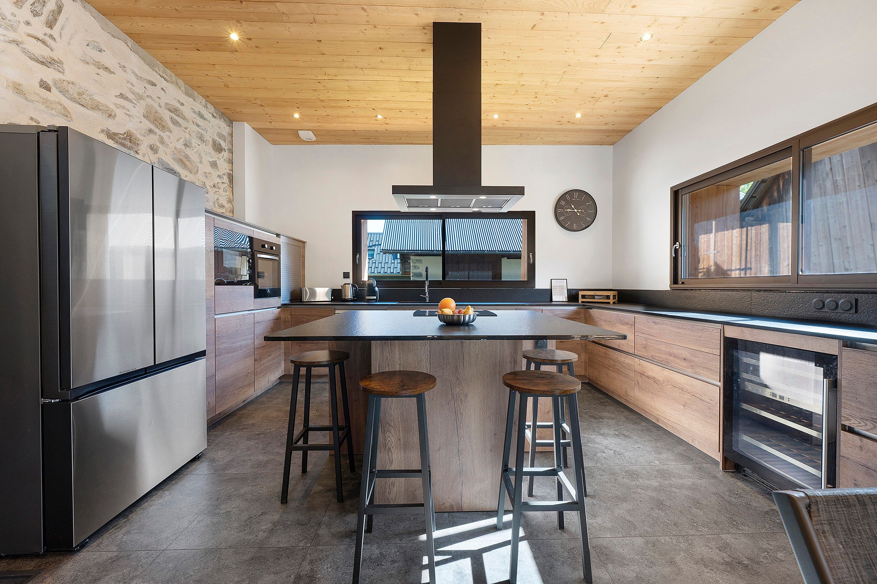 A modern kitchen with a central island, bar stools, stainless steel appliances, and a large window.