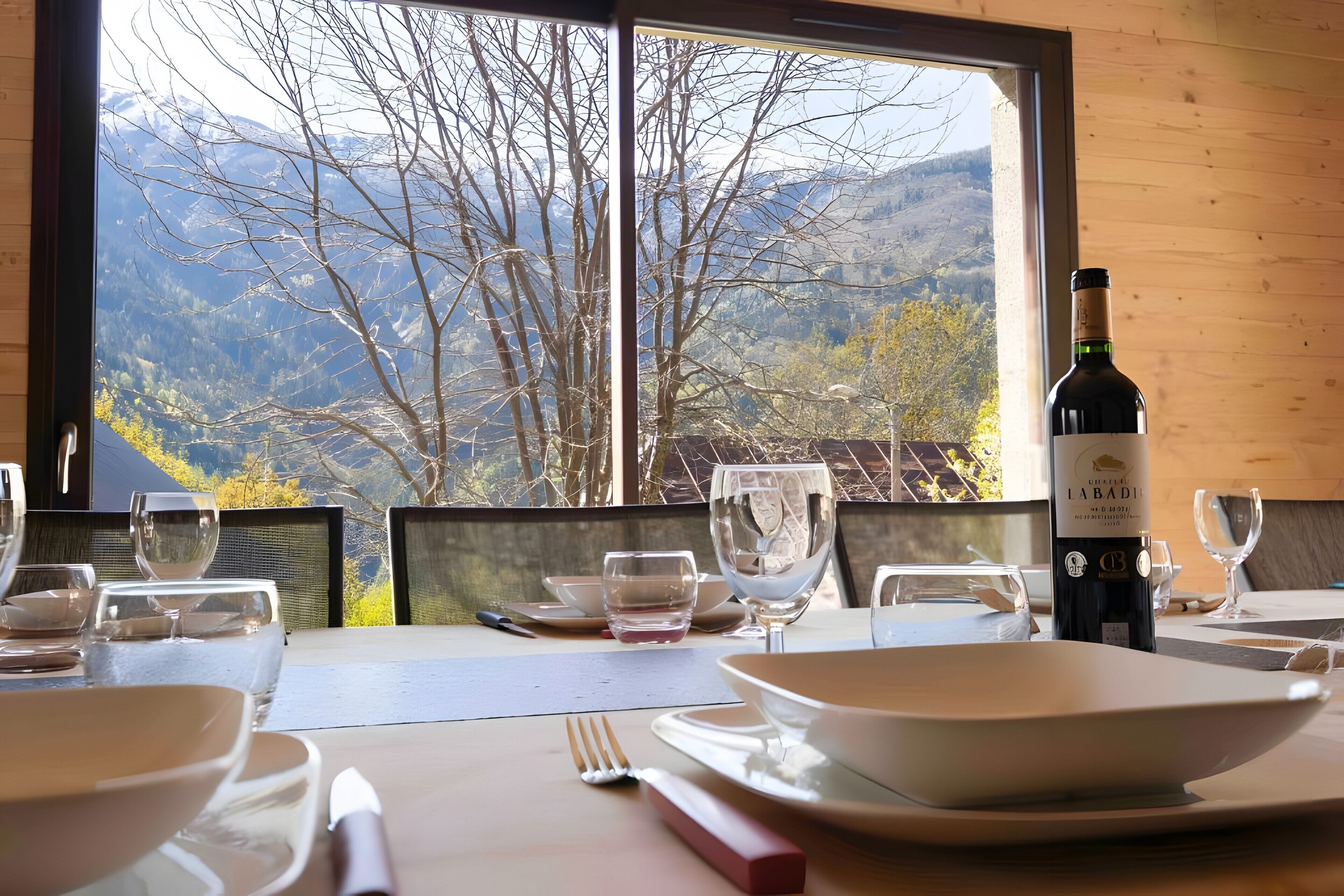 A dining table set with plates, glasses, and cutlery, with a bottle of wine and a view of mountains through a window.