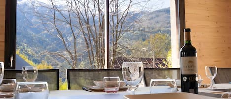 Mesa de comedor preparada con platos, vasos y cubiertos, con una botella de vino y vistas a las montañas a través de una ventana.