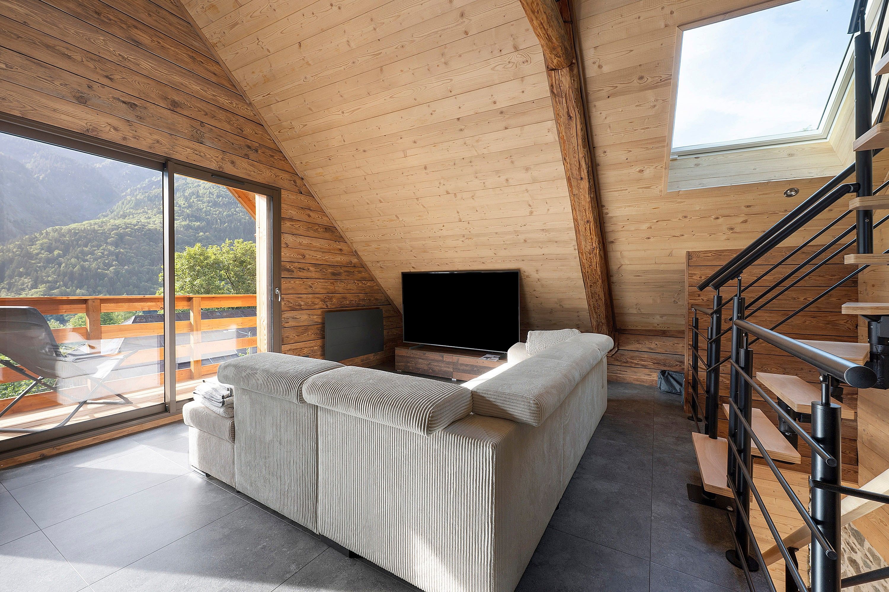 A cozy, sloped-roofed room with a large window, a flat-screen TV, a sofa, and a staircase.