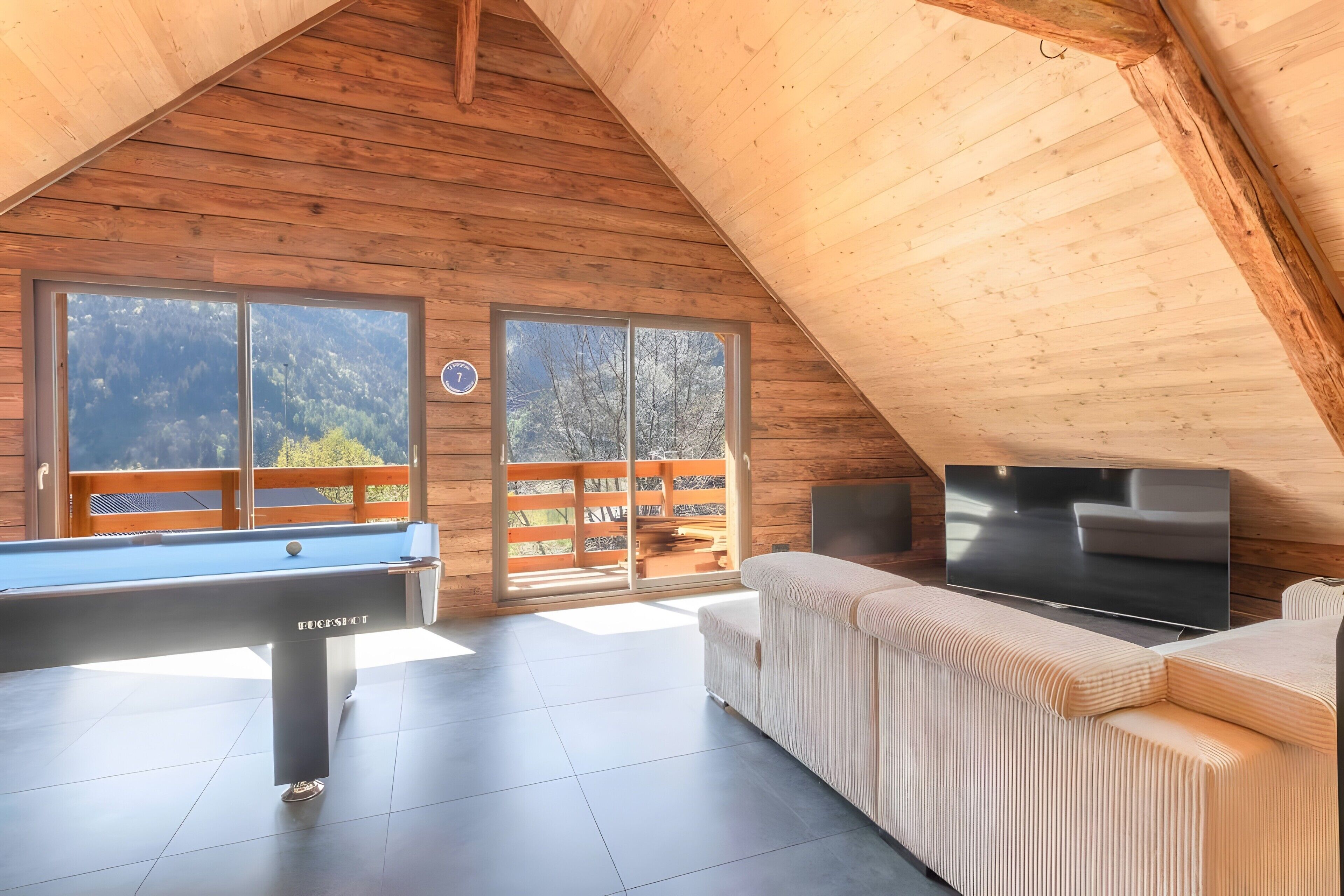 A wooden interior with a pool table, a sofa, a flat-screen TV, and large windows offering a mountain view.