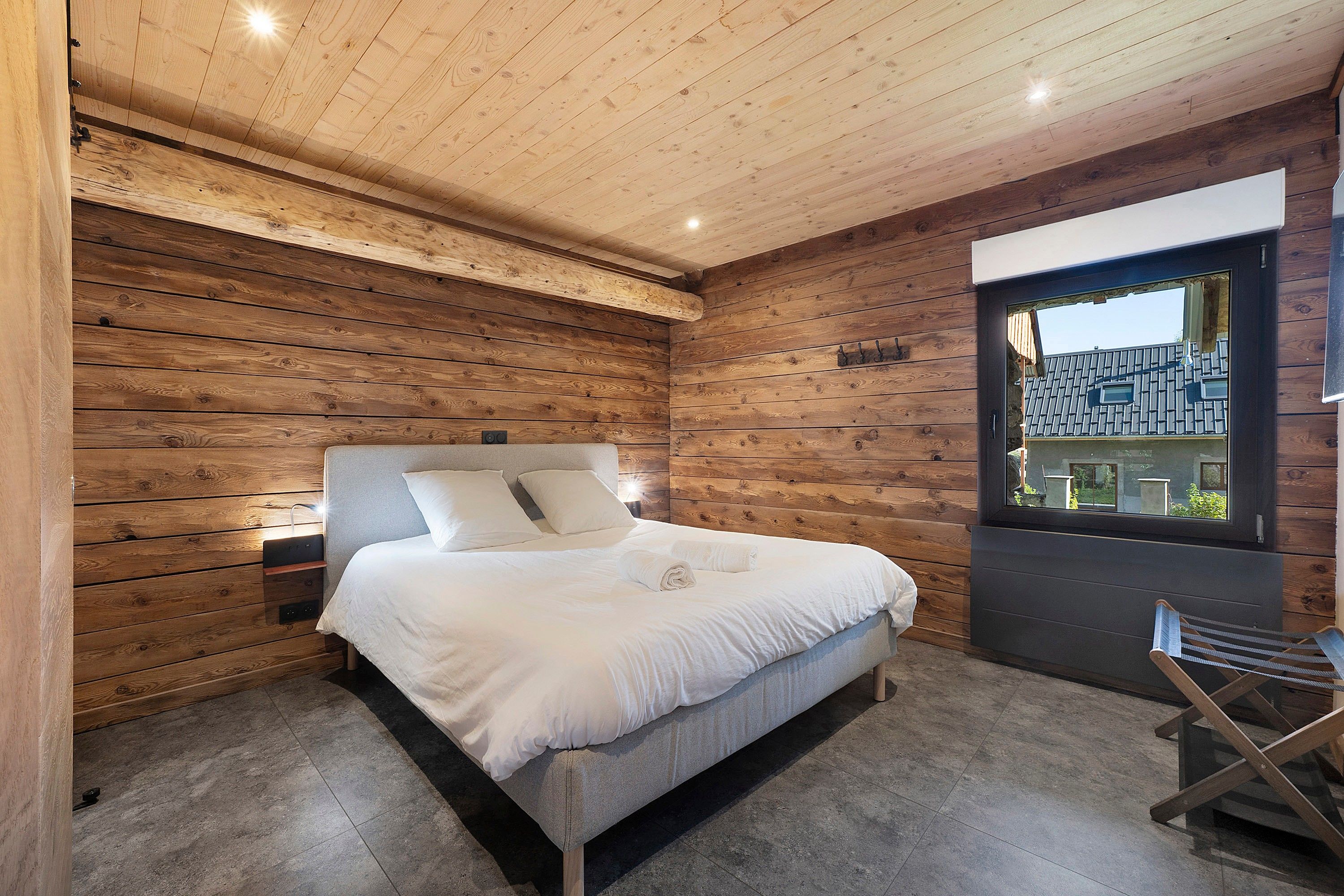 Une chambre avec un plafond en bois, un lit avec une literie blanche, une table de chevet et une fenêtre donnant sur une maison.