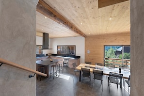 A modern kitchen with a central island, wooden ceiling beams, and a dining area with a view of greenery outside.