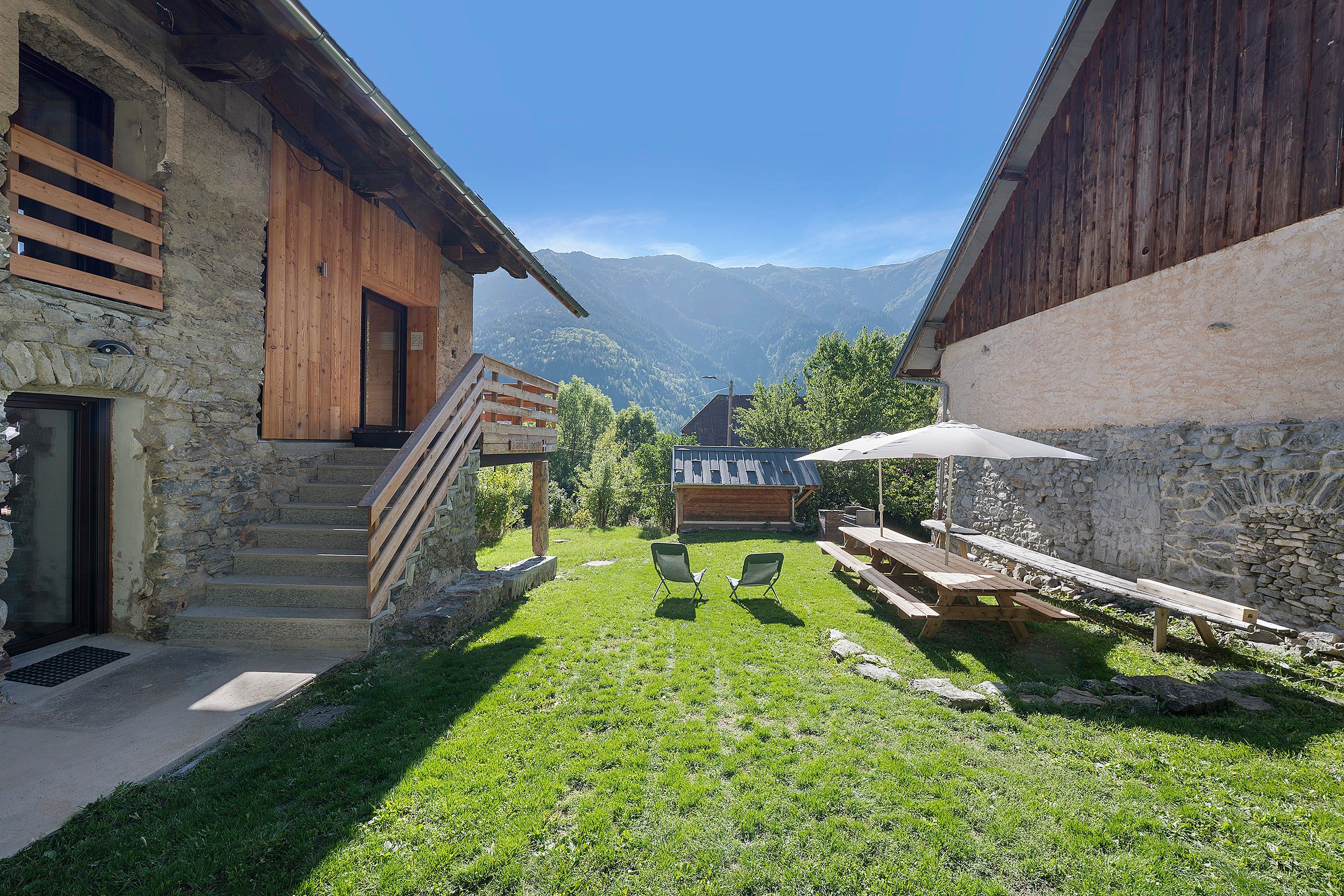 A rustic outdoor area with a wooden staircase leading to a wooden door, a picnic table with chairs, and a stone wall.