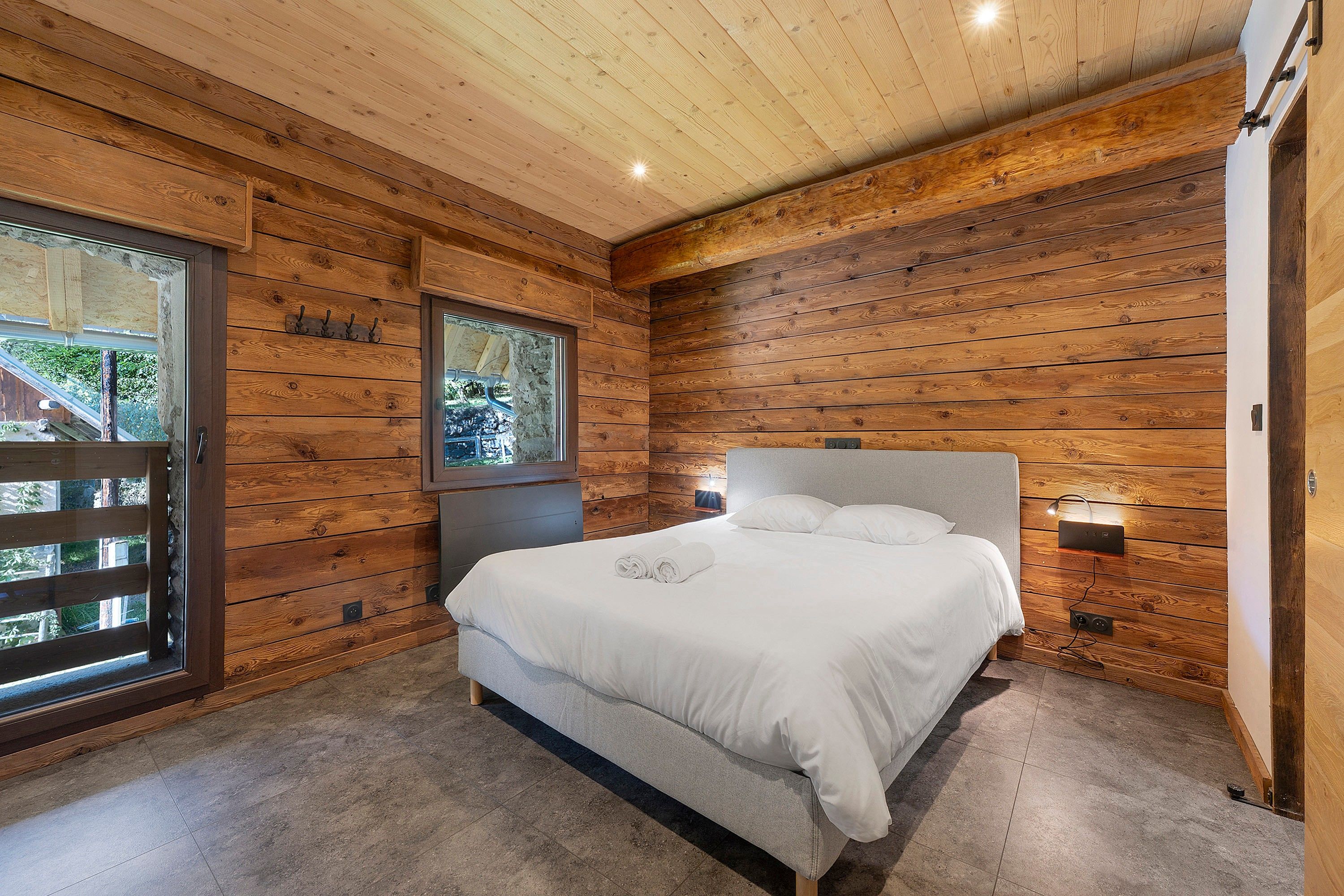 Un dormitorio con paneles de madera, una cama, un ventanal e iluminación en la pared.
