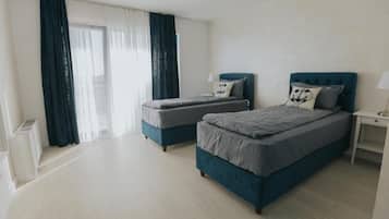 Quarto de hotel com duas camas, um radiador branco, uma mesa de cabeceira e uma janela com cortinas.