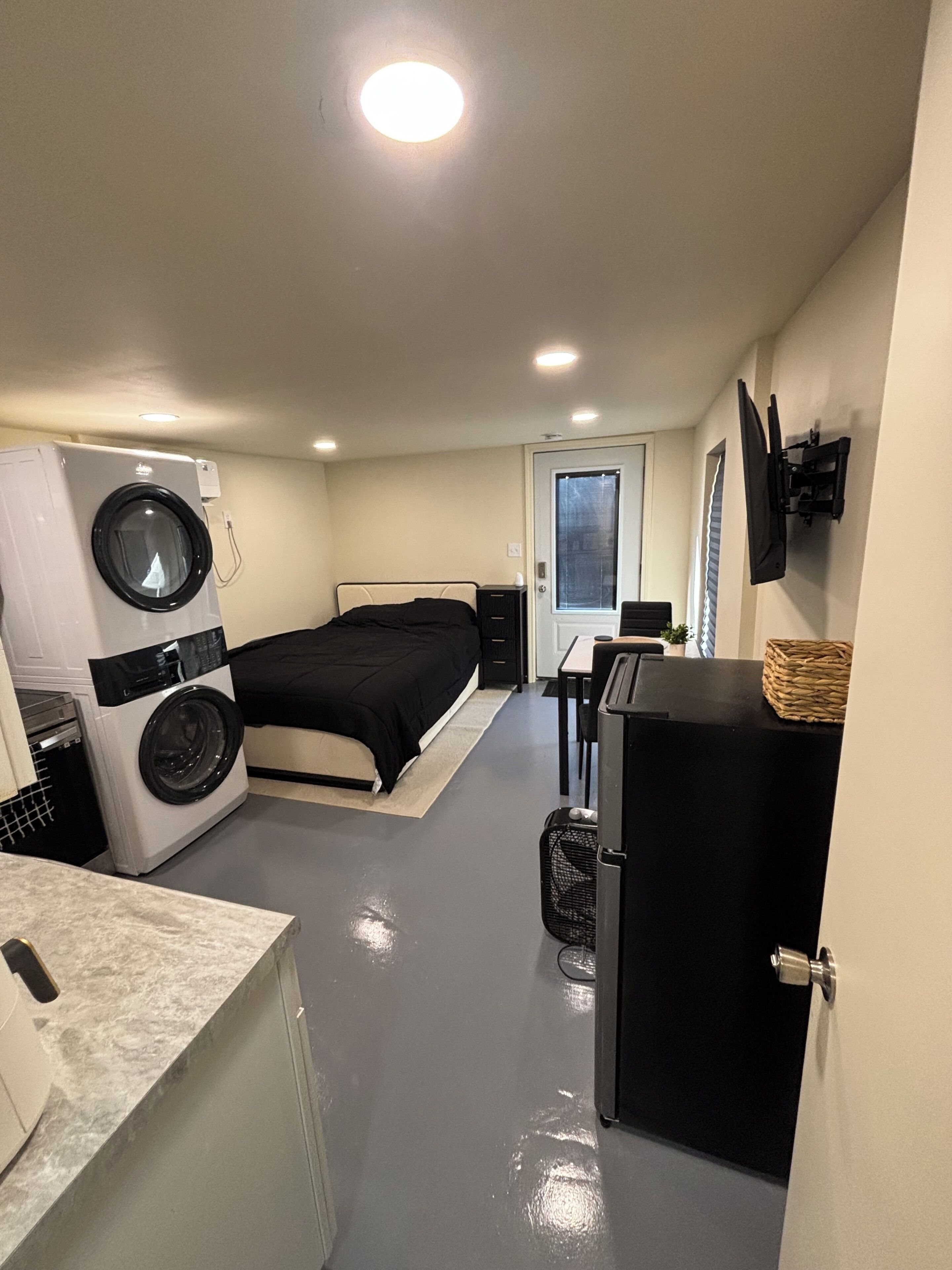 A compact hotel room with a bed, a washing machine, a small kitchenette, and a TV mounted on the wall.