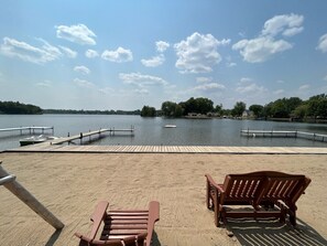A sandy beach with two wooden Adirondack chairs and a wooden dock extending into a calm lake.