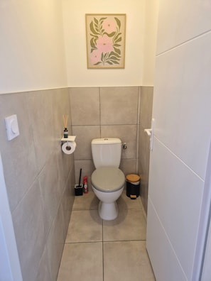A small bathroom with a toilet, a trash bin, a toilet paper holder, and a decorative wall piece. - The Catalan Cocoon – 3 bedrooms, garden & barbecue – sleeps 6 (Perpignan)