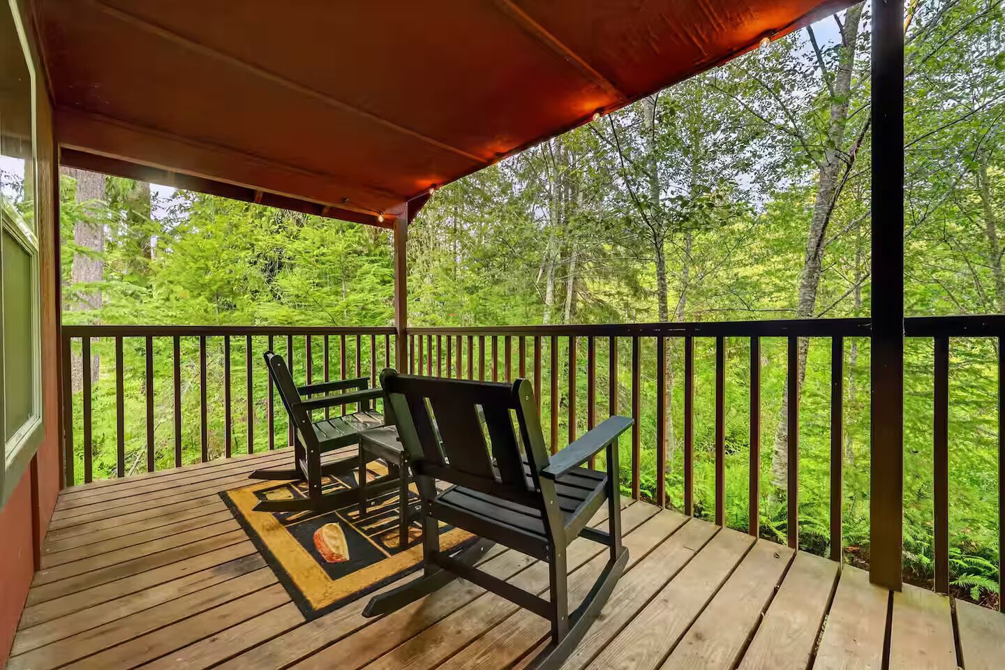 Una terraza de madera con dos mecedoras y una mesa, con vistas a un área boscosa.
