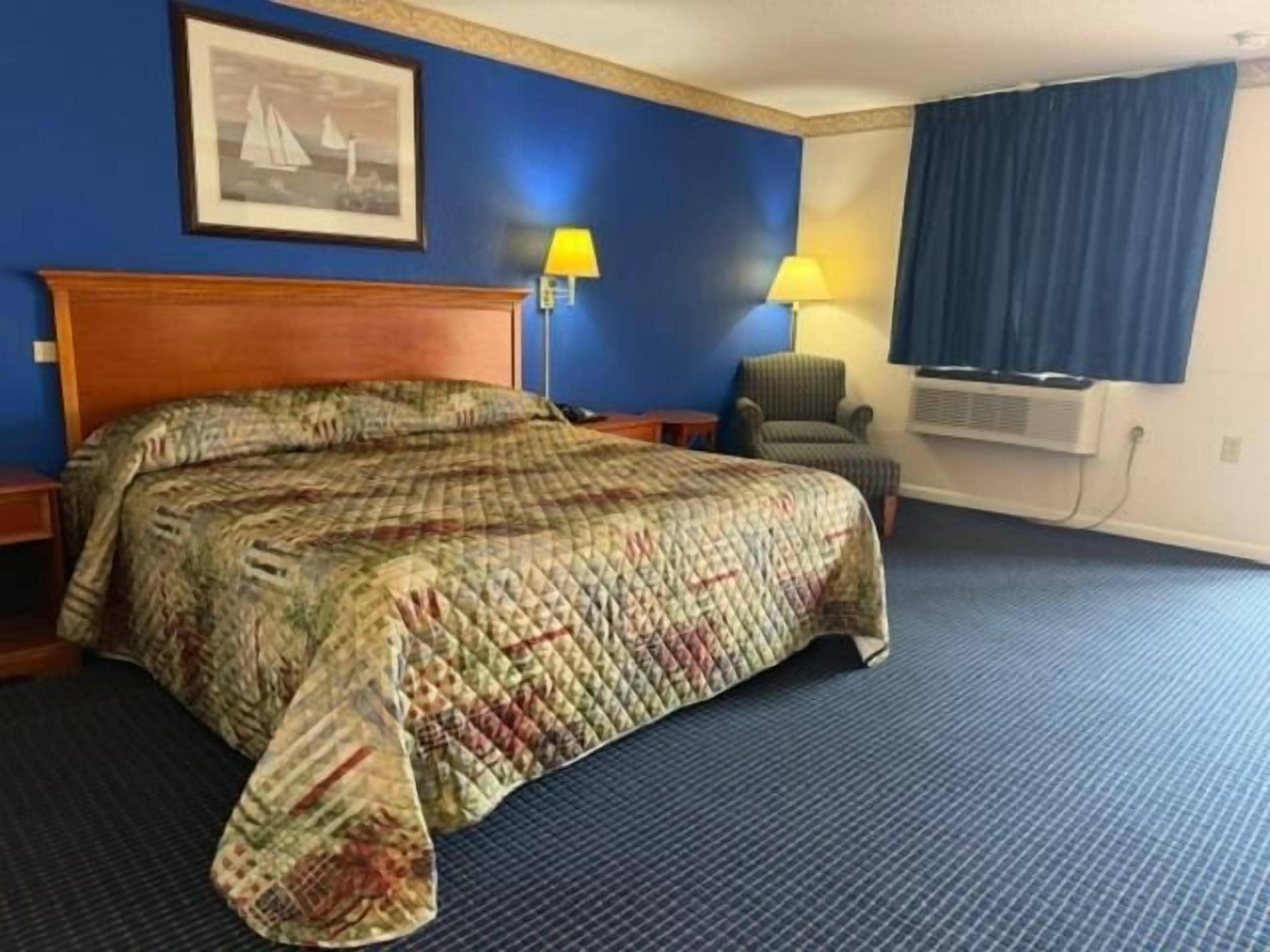 A hotel room with a large bed, a chair, a desk, and a wall-mounted air conditioner.