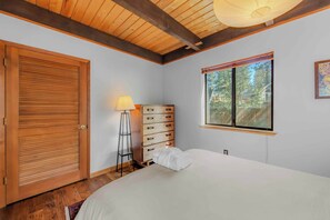 A bedroom with a wooden door, a dresser with drawers, a bedside lamp, a window with a view of trees, and a bed with a white comforter. - Lakewood Escape 3BR Retreat (South Lake Tahoe)
