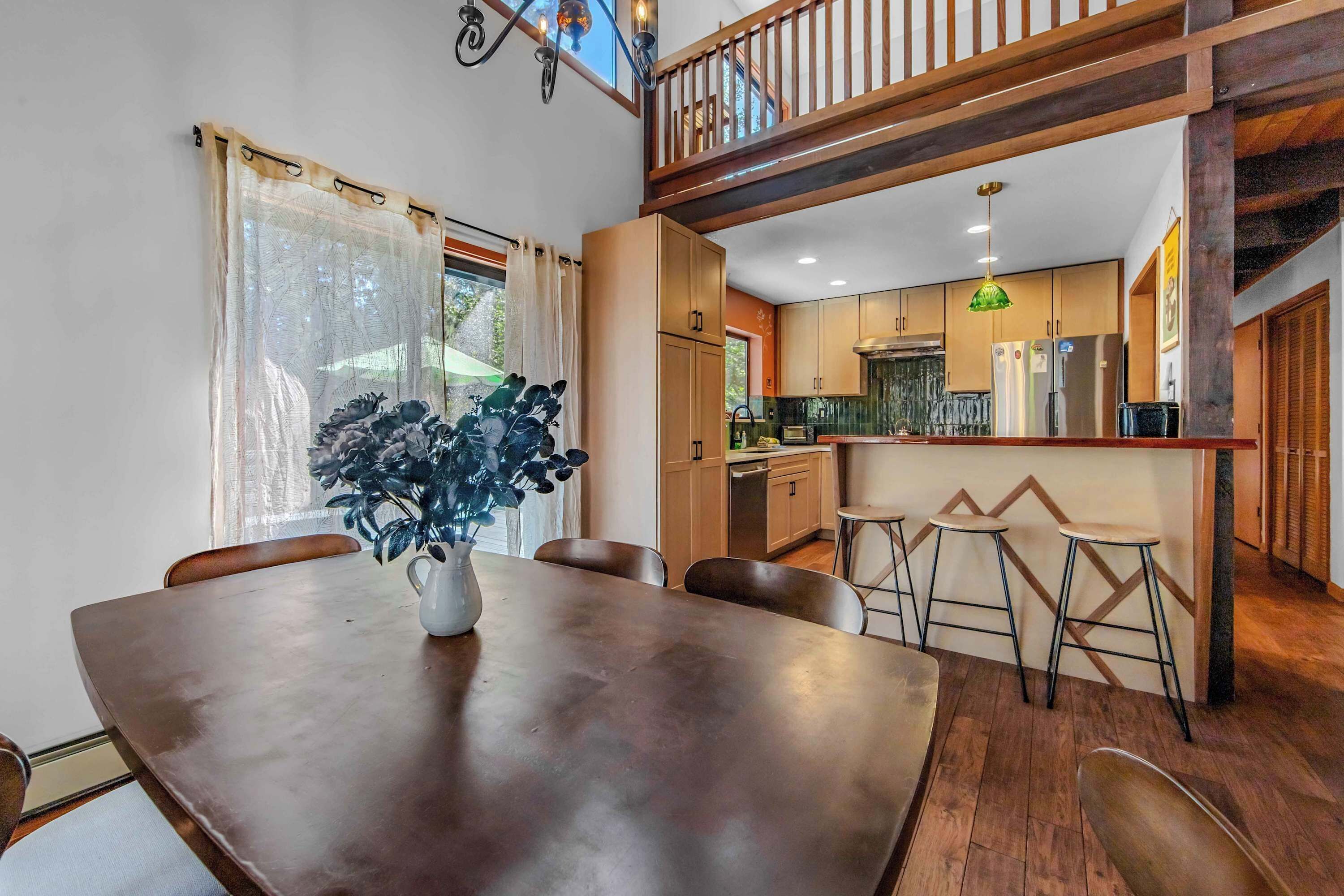A dining area with a wooden table and chairs, a vase with flowers, and a view of the kitchen and upper floor.