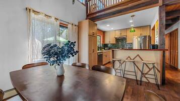 A dining area with a wooden table and chairs, a vase with flowers, and a view of the kitchen and upper floor.
