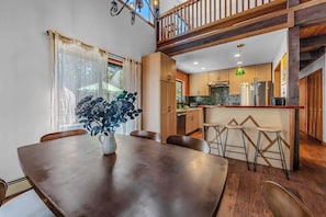 A dining area with a wooden table and chairs, a vase with flowers, and a view of the kitchen and upper floor.