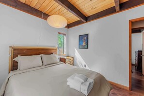 Ein Schlafzimmer mit Holzboden, einem Bett mit Kopfteil, einem Nachttisch, einem Fenster mit Blick auf Grünflächen und einem gerahmten Bild an der Wand.