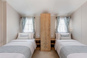 A bedroom with two beds, a central wooden wardrobe, and curtains on the windows. - Host & Stay | Meadow View (Winchelsea)