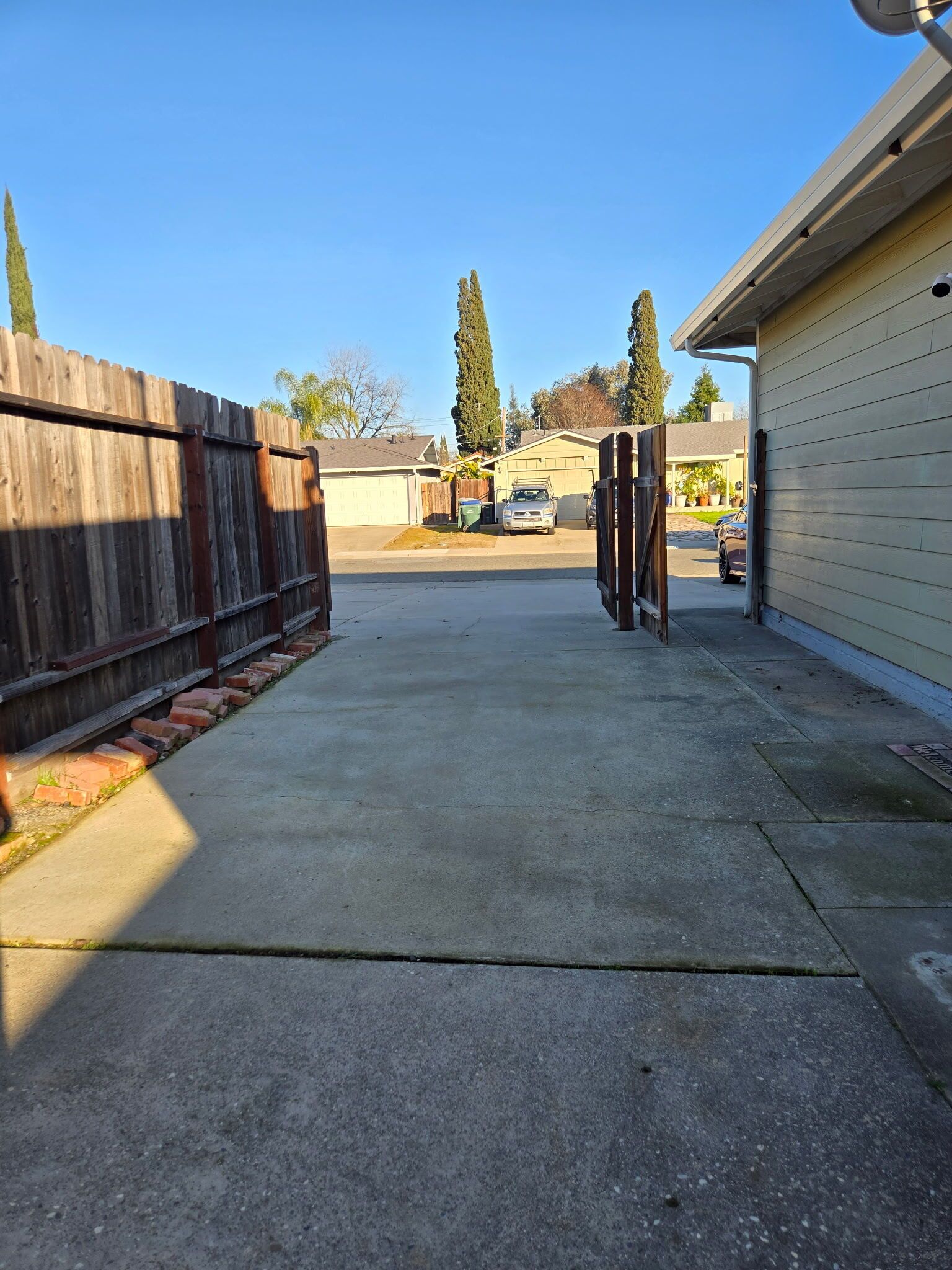 Une allée en béton menant à un garage, avec une clôture en bois d’un côté et un bâtiment de l’autre.