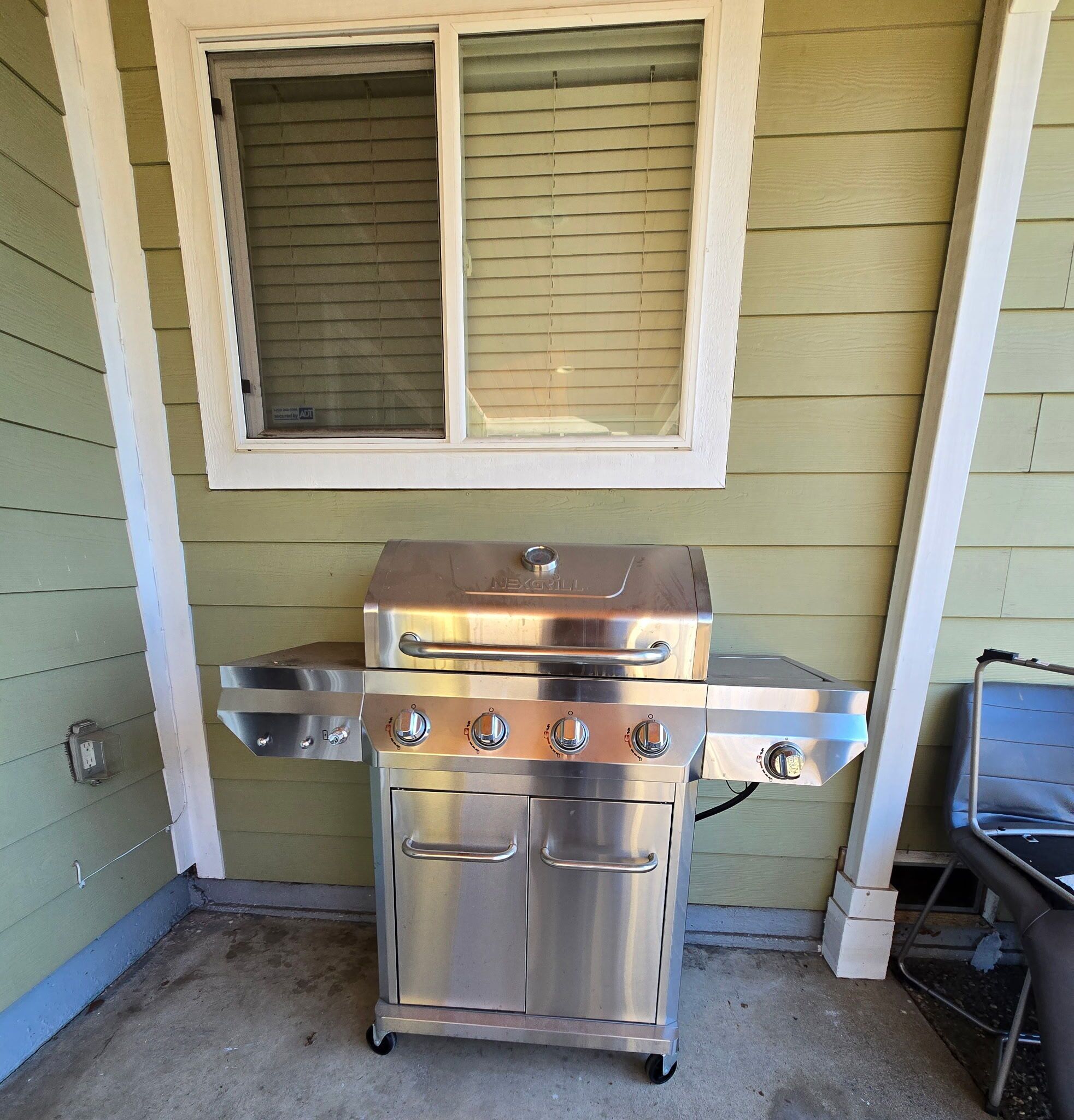 A stainless steel outdoor grill with four burners, a side burner, and a storage cabinet, positioned under a window with blinds.