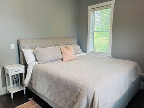 A bedroom with a bed, a nightstand, a window with blinds, and a view of greenery outside. - 3 King Beds Minnehaha Hideaway Near Downtown (Minneapolis)