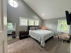 Quarto com teto inclinado, uma cama com cabeceira, duas mesas de cabeceira, uma cadeira e uma janela grande com persianas.