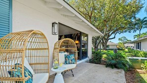 A covered outdoor seating area with wicker chairs and a table, a house entrance, and a well-maintained garden. - PGA Resort Home – Heated Pool, Walk to Dining & 15 min Drive to Beaches (Palm Beach Gardens)