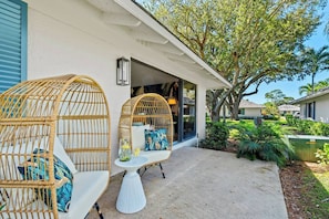 A covered outdoor seating area with wicker chairs and a table, a house entrance, and a well-maintained garden. - PGA Resort Home – Heated Pool, Walk to Dining & 15 min Drive to Beaches (Palm Beach Gardens)