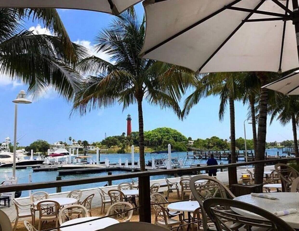 An outdoor dining area with tables and chairs, overlooking a marina with boats and a lighthouse.