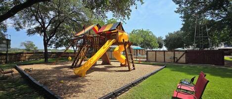 A playground with a slide and climbing structures, surrounded by a fenced area and trees.