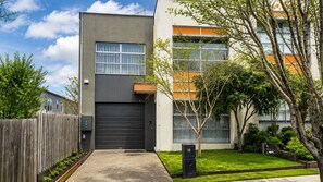 A modern two-story residential building with a dark grey facade, a large garage door, and a small tree in the front yard.